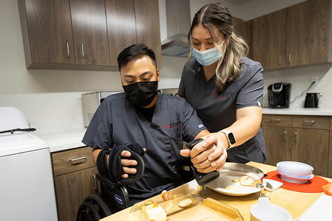 Two occupations therapy students working through a simulation in a kitchen. One is assisting the other that has limited use of their hands.