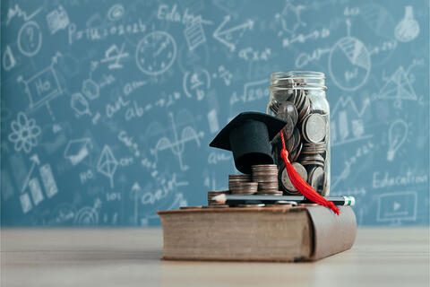 A jar of coins and a graduation cap on top of a book with a blackboard in the background