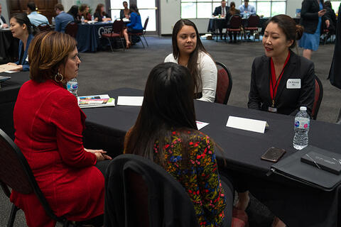 Four people sitting around a table, two on each side. In the background there are other tables with other people.