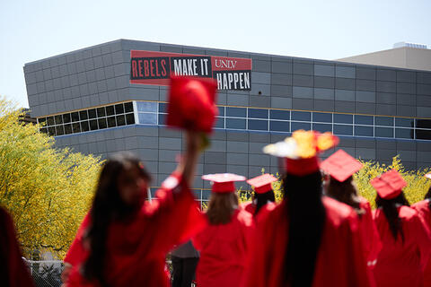 eight UNLV graduates walking in their cap and gowns with the sign "rebels make it happen" afar