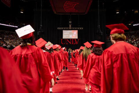 A rear view of UNLV students, dressed in red caps and gowns, filtering into the Thomas &amp; Mack Center with the stage in the background