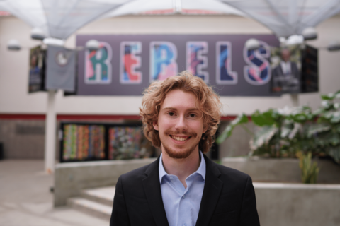 Sky Denson in shirt and open blazer in front of Rebels sign