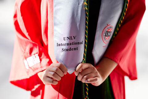 Close-up of a graduate wearing a red gown and holding a silver graduation stole that reads "UNLV International Student."