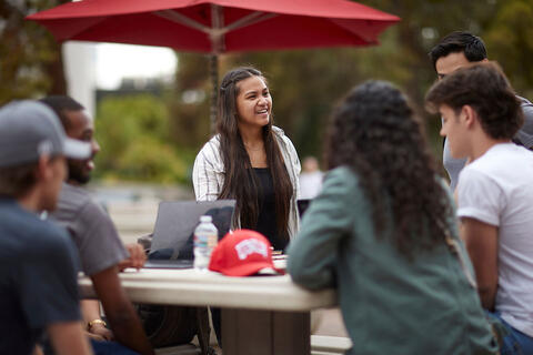 A group of students sitting around an outdoor table under a red umbrella, talking and smiling.