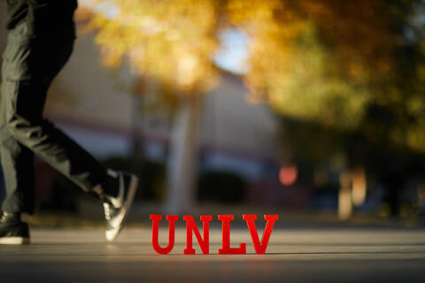 the lower half of a person is visible as they walk by UNLV letters on the sidewalk