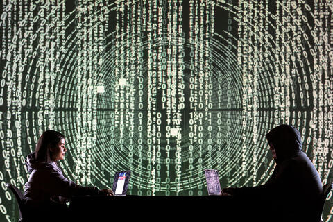 Two individuals working on laptops in front of a large digital screen filled with binary code.