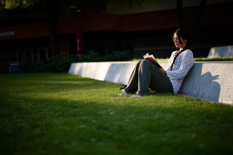 A woman sits down on grass leaning against a concrete ledge while reading a book.