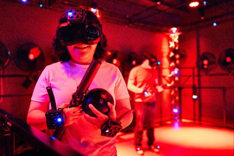 Female student in white shirt wearing a virtual reality headset and hand trackers smiles while enjoying the free roam pod inside UNLV's Dreamscape Learn