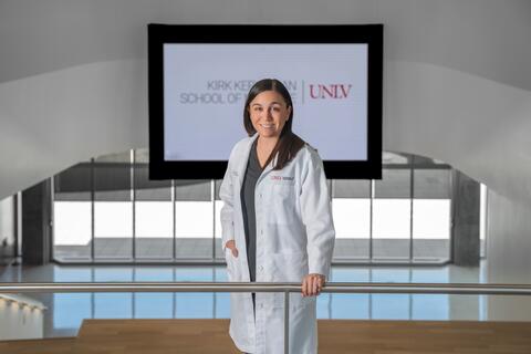 Woman in white medical coat with the UNLV School of Medicine logo poses in front of a UNLV School of Medicine sign with her right hand in her pocket and left hand on a silver rail
