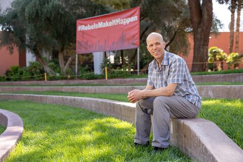 Bald white man in plaid shirt and gray pants smiles while sitting on a short wall outdoors on UNLV's campus