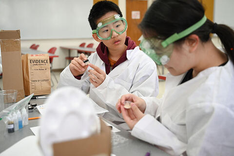 Two students in lab coats and goggles working on a science experiment in a classroom.