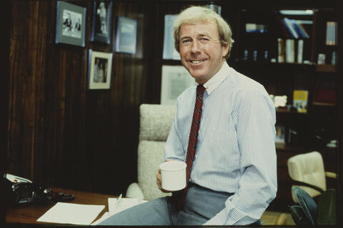 archive photo of man in tie holding coffee cup and leaning on office desk