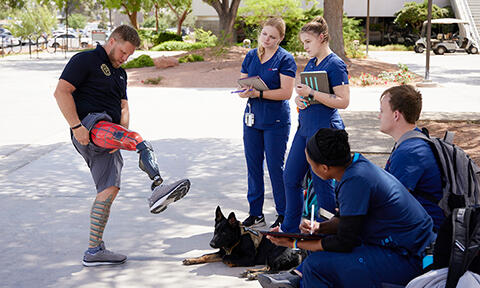 A man showcases a lower-limb prosthetic while standing outdoors as a small group of nursing students in blue scrubs observe and take notes.
