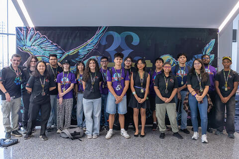 A group of students and mentors standing indoors in front of a large, colorful event backdrop
