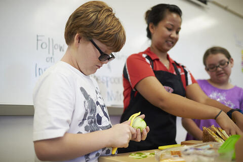 A student peels an apple while two people in the background assemble sandwiches.
