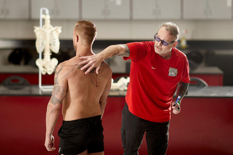 An instructor demonstrates back anatomy by pointing to a student’s shoulder and upper back in a classroom.