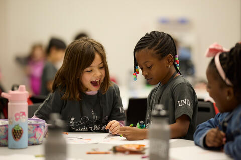 Three young girls excitedly watch as one draws on paper with a crayon