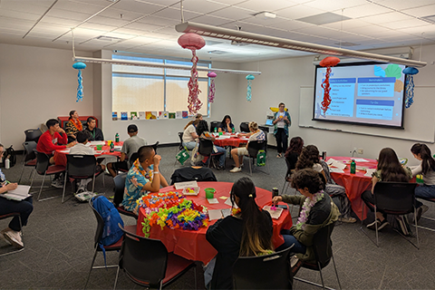 People sitting around round red tables as a presenter speaks with a slideshow behind them