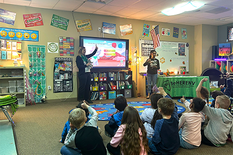An instructor standing in front of a TV with students sitting on the floor holding their thumbs up or down