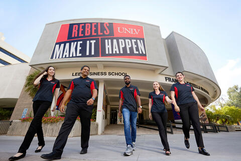 Five students wearing black and red polo shirts walk in front of the Frank and Estella Beam Hall beneath a large “Rebels Make It Happen” sign.