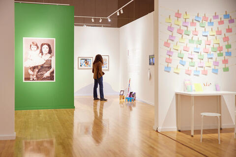 Interior view of an art gallery with wooden floors and white walls. A visitor stands facing framed artworks in the center of the space.