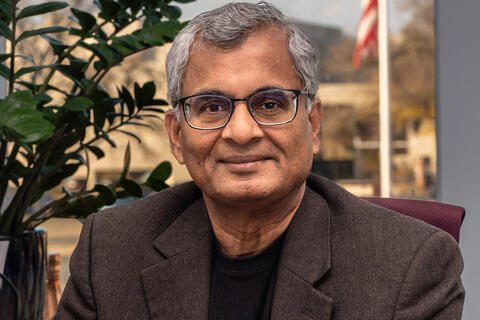 man in jacket posing iman in jacket posing in an office with image of buildings and flag visible behind himn an office with buildings and flag visible through window.