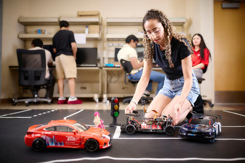 A student kneeling on the ground with 3 vehicles and a robotic car laying on a black mat with white lines