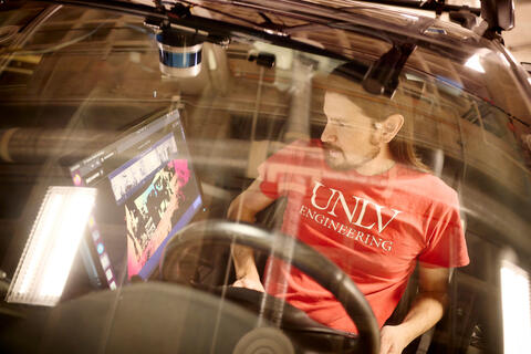 A man in a red U-N-L-V t-shirt looking at a computer in a car