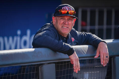 man in UNLV baseball cap and gear leaning over a dugout fence