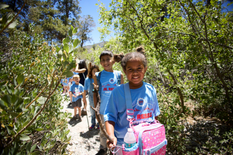 A line of children in blue t-shirts walking through a clear pebble path with a wall of trees on each side