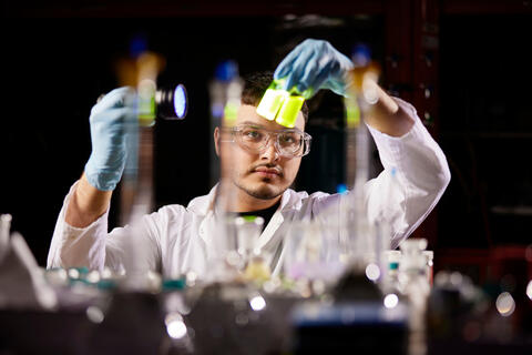 A man in a white coat with blue gloves holding two bottles in his hands with a flashlight pointed at the bottles