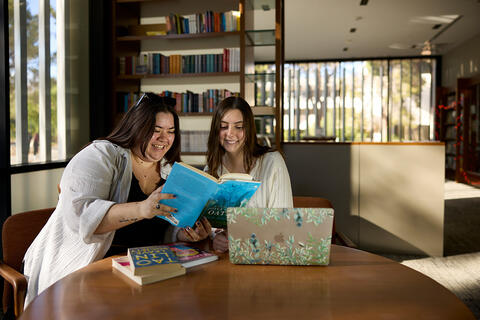 Two students sit together at a table, reading a book