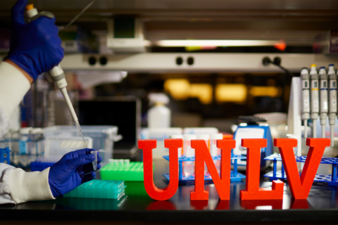 A pair of hands in blue latex gloves holding a test tube and a needle with a wooden sign on the table that says "U-N-L-V"