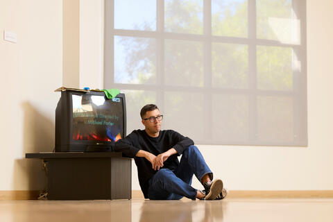 Michael Porter sits next to a TV, part of the 'On Time' exhibit