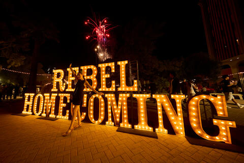 woman posing in front of outdoor display saying "Rebel Homecoming" in lights at night.