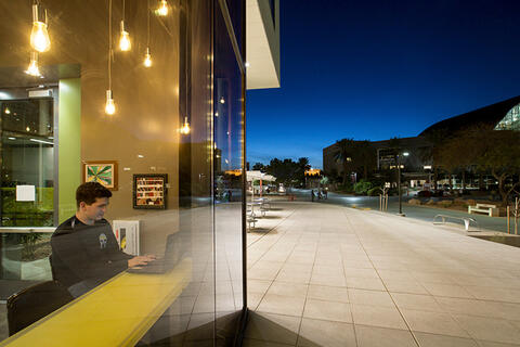 A man working on a laptop at night looking out a window at the campus outside.