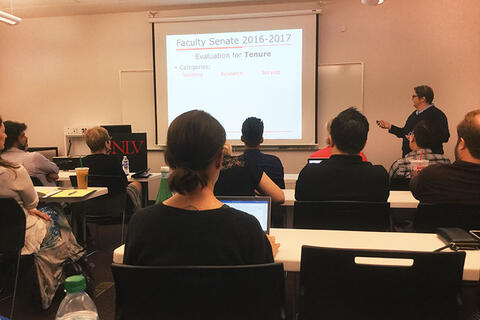 Woman presenting a slideshow about tenure to a group of people sitting down behind white tables