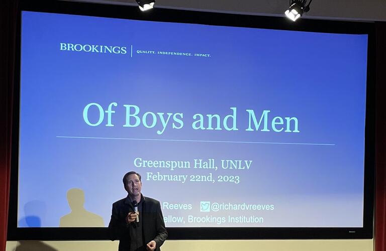 Richard V. Reeves delivering his &quot;Of Boys and Men&quot; lecture on February 22. He stands before a large screen displaying the name of his lecture on a blue PowerPoint background.