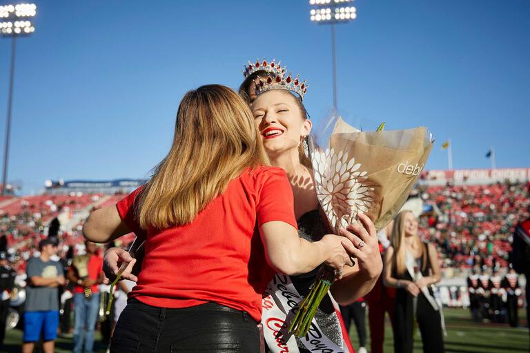A student hugs the UNLV president while holding a bouquet and wearing a crown at a football game.