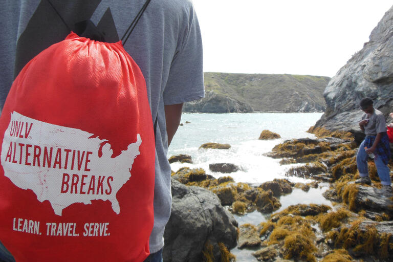 student with red Alternative Breaks backpack facing a rocky ocean coast