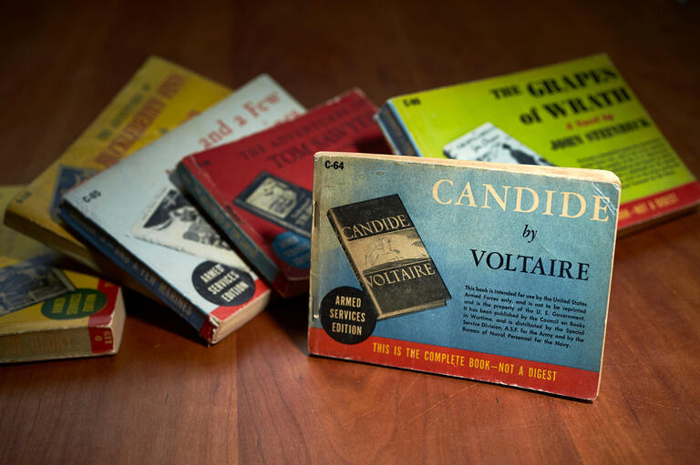 A pile of books is topped by a special edition of Candide by Voltaire.