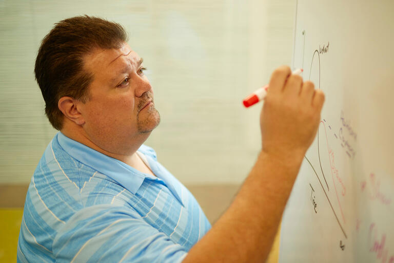 A man in a blue striped shirt writes on a whiteboard.
