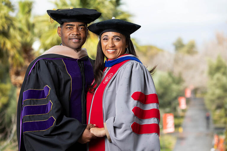 Keith Rogers and Rebecca Rogers, wearing graduation robes, pose overlooking a pathway on the UNLV campus.
