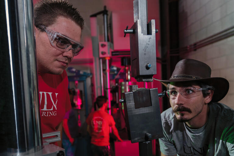 A UNLV student wearing goggles, and one wearing a hat and glasses, stare at materials in a vice.
