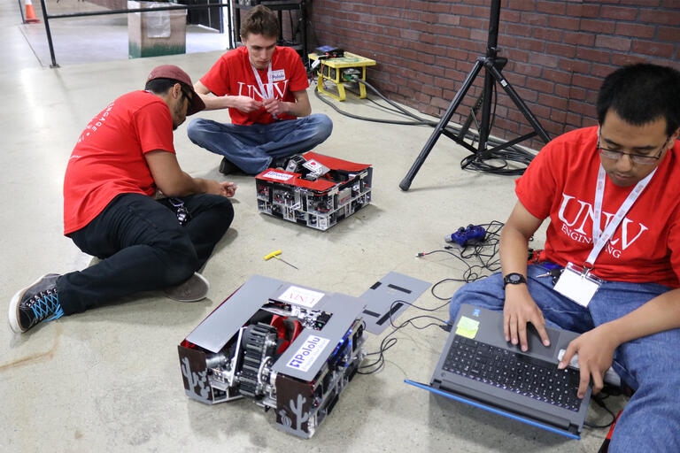 Engineering students use a laptop and tools to fix two box-like robots