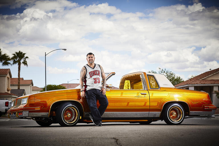 A man in a UNLV basketball jersey leans on a gold-colored Oldsmobile.