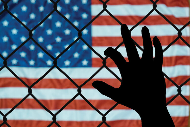 A hand reaches up to a chain link fence in front of the American flag.