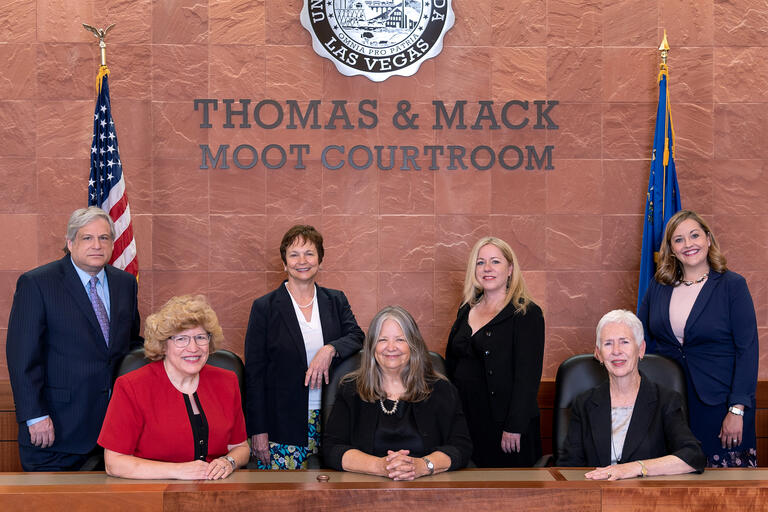 Seven law professors pose at the bench in the Thomas &amp; Mack Moot Courtroom between the United States and Nevada flags.