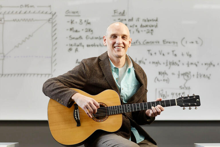 A man sits in front of a whiteboard holding an acoustic guitar