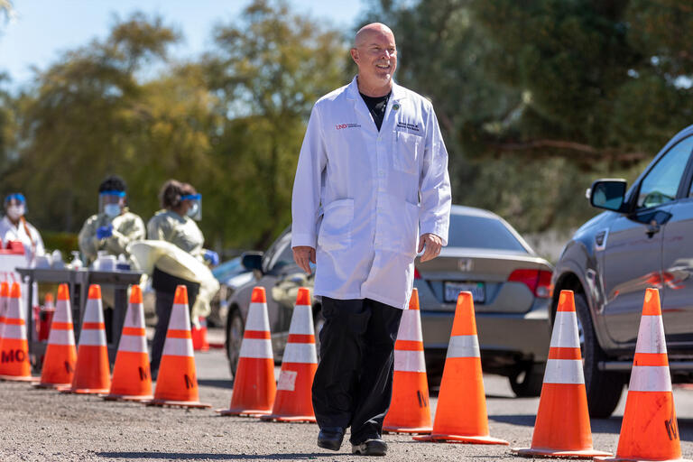 A doctor in a white lab coat walks past a row of orange cones and a line of cars.